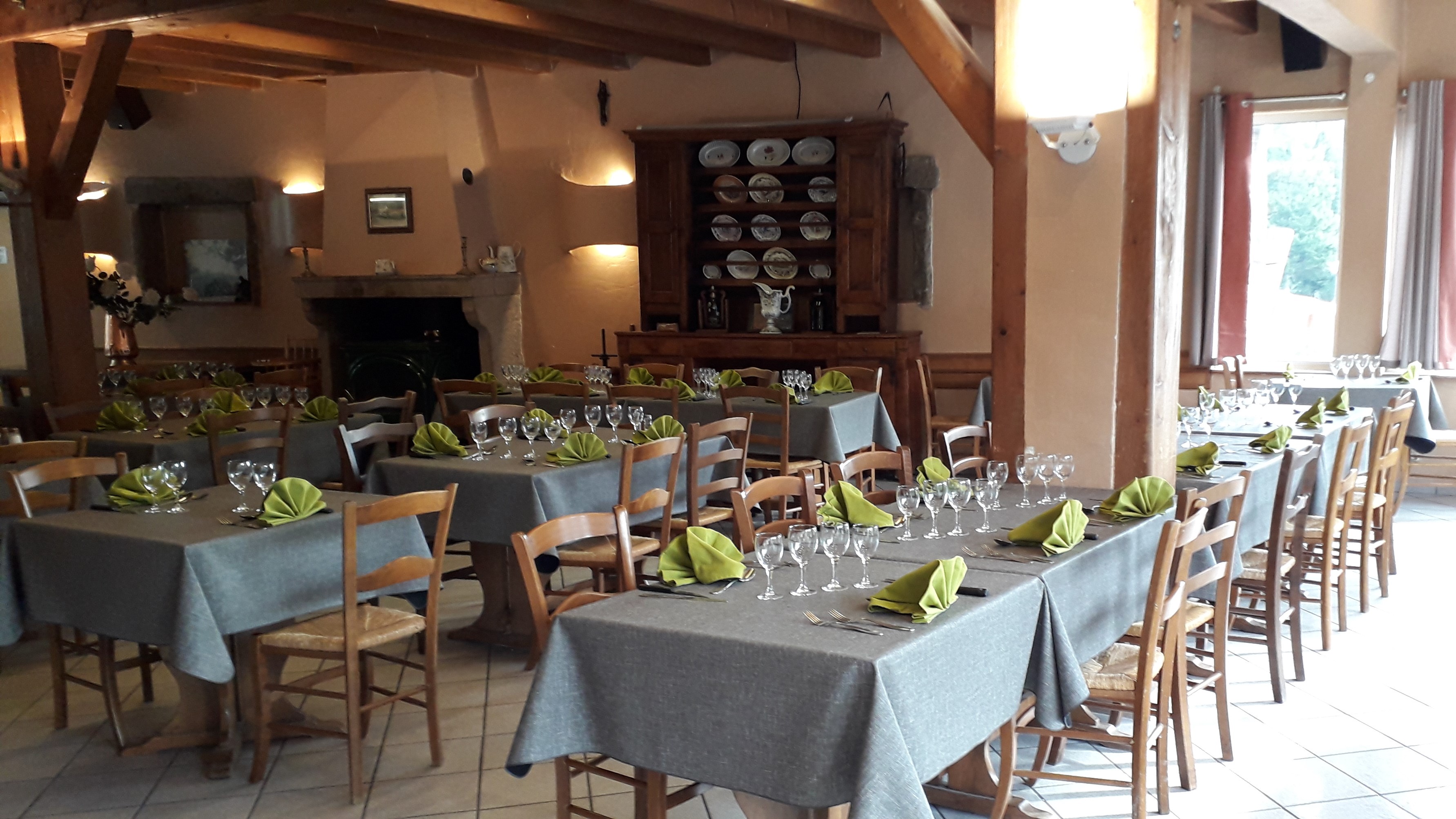 Salle de restaurant de l’Auberge du Grand Bois à La Versanne, tables dressées avec nappes grises et serviettes vertes, poutres en bois et cheminée. Photo issue de la fiche de l’Office de Tourisme du Pilat sur Apidae.